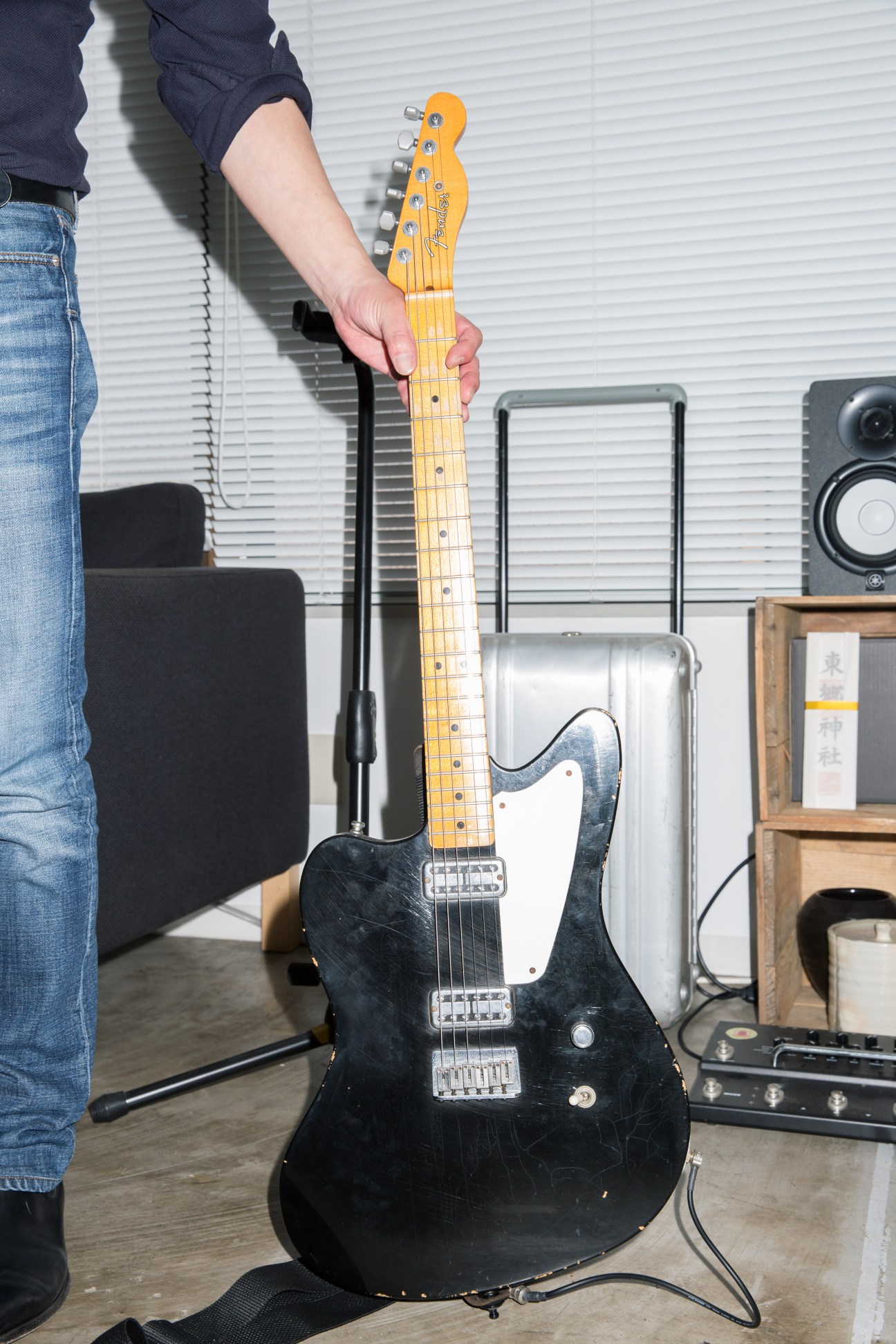 Person holding a black electric guitar with yellow maple neck in a music studio setting.