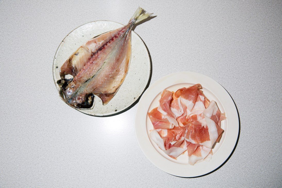 Dried salted fish on a plate and sliced prosciutto on another plate against a white background.