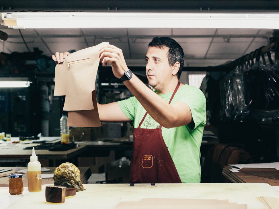 Leather craftsman inspecting a tan leather pouch at a factory workbench in Ubrique.