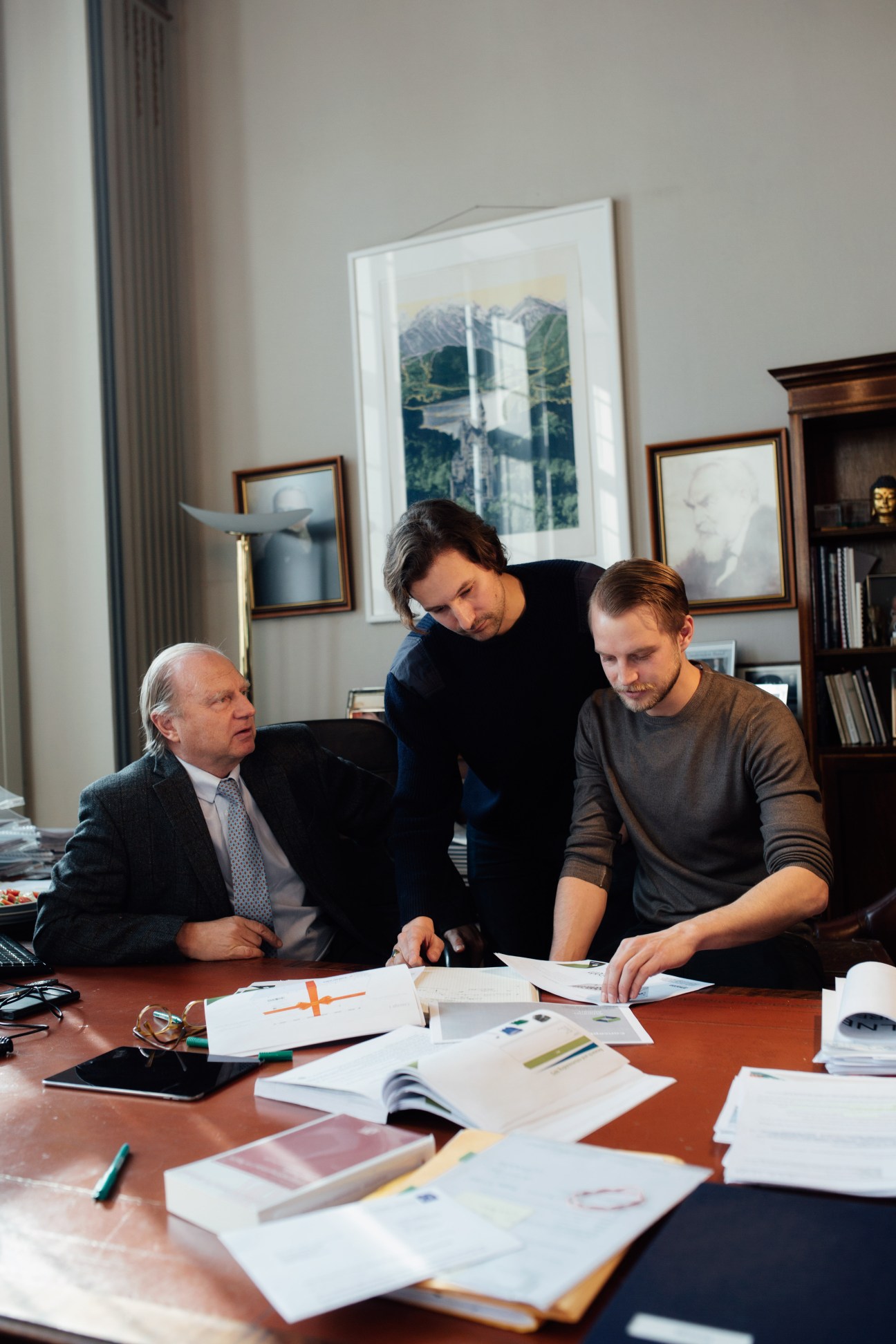 Three men review documents at a wooden table in an office with framed artwork on the wall.