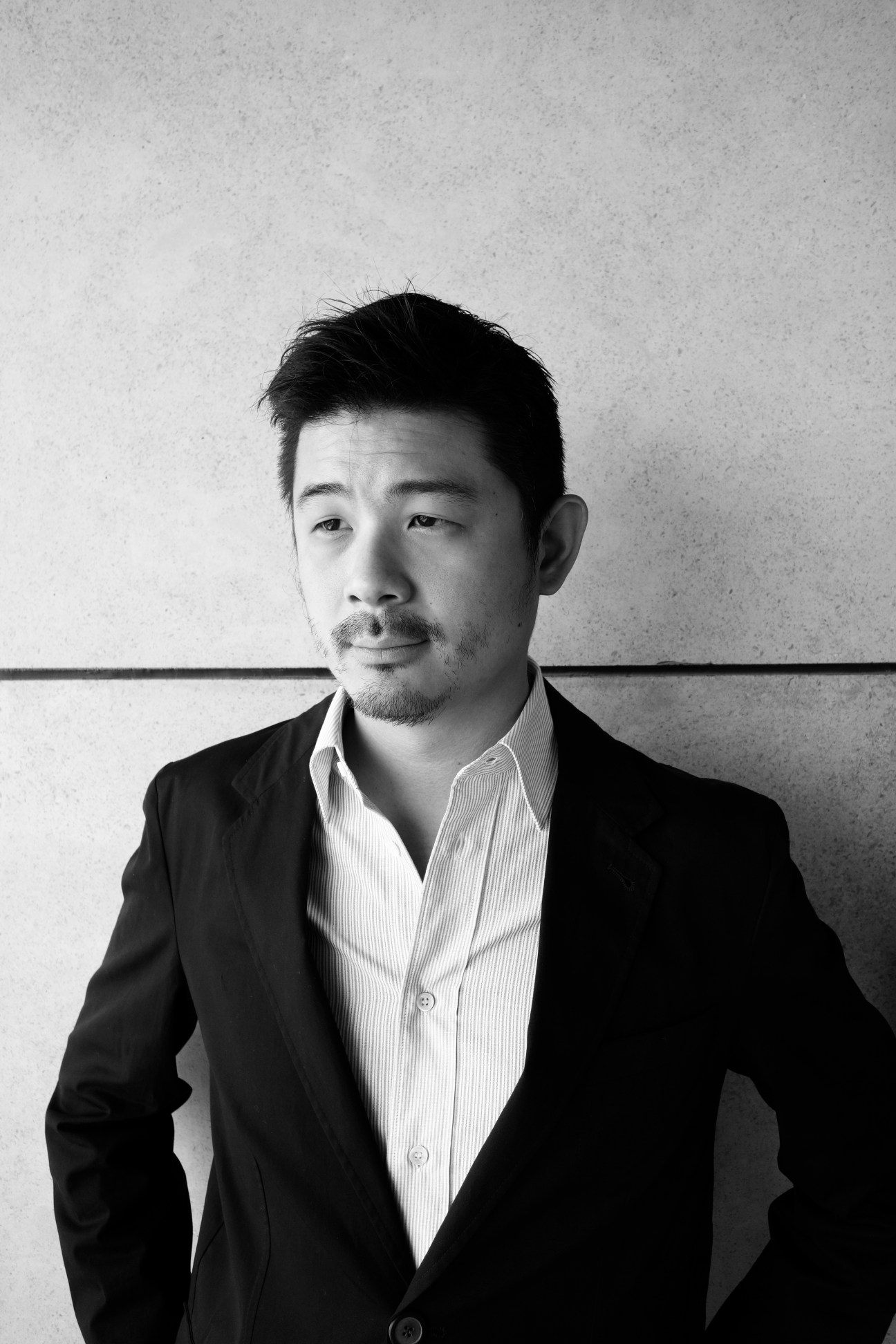 Black and white portrait of an Asian man wearing a dark blazer and white striped shirt against a textured wall.