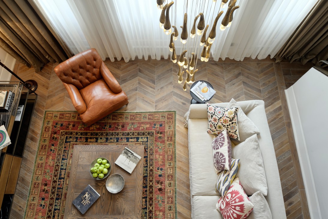 Overhead view of a modern hotel room with herringbone wooden flooring, cream sofa with patterned pillows, cognac leather armchair, ornate rug, and brass hanging pendant lights.