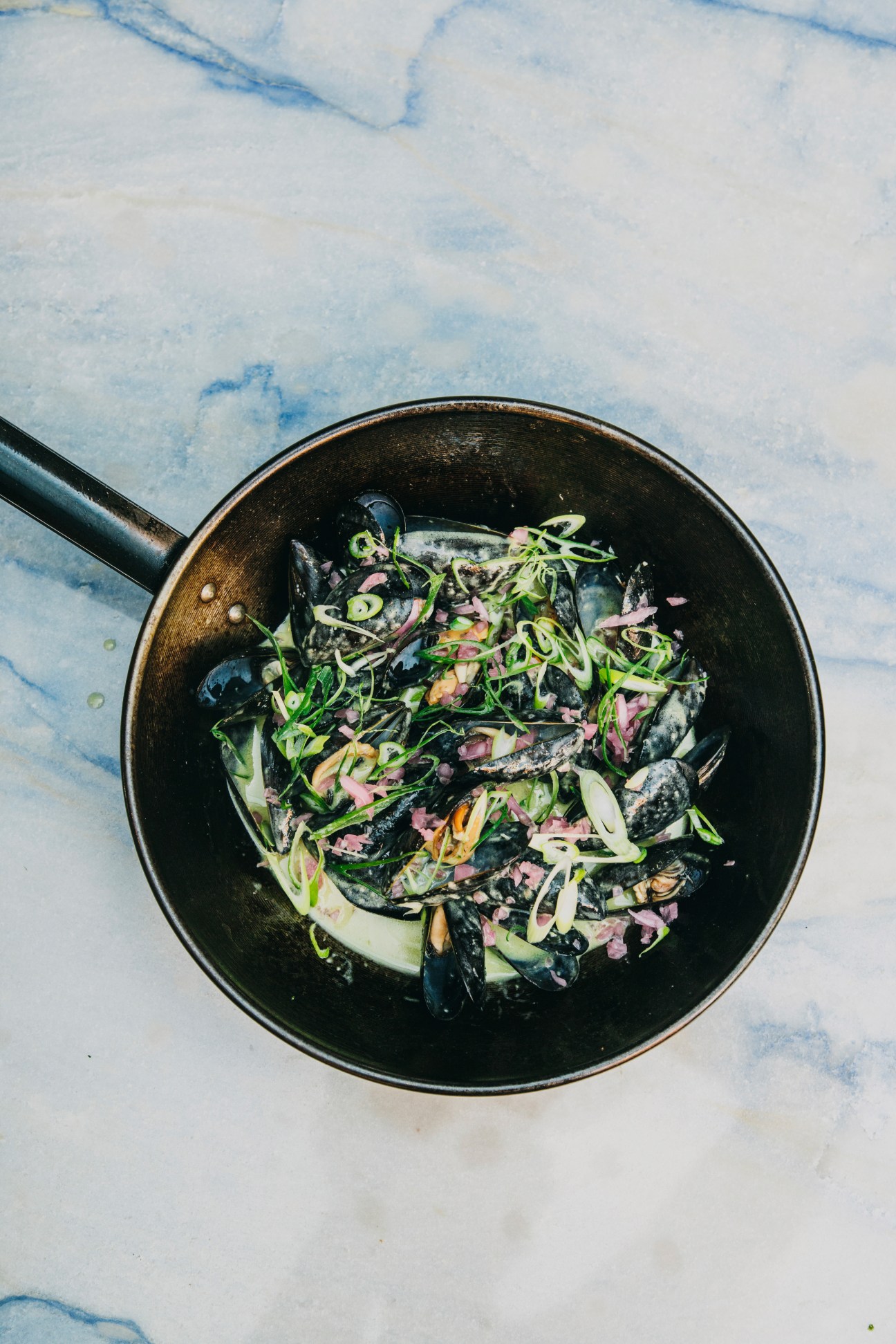 Green curried mussels in a black bowl garnished with fresh chives and herbs on a marble surface.