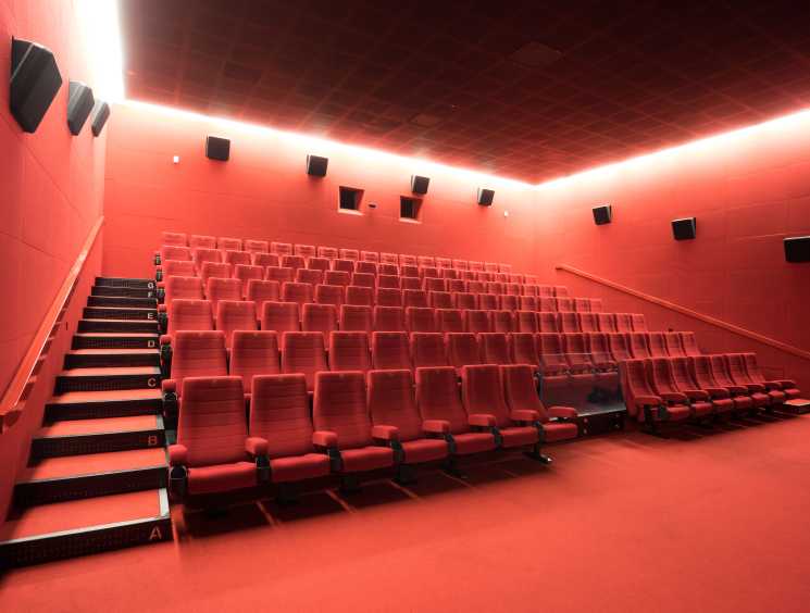 Modern cinema auditorium with red seats and walls, linear ceiling lights, and labeled seating rows.