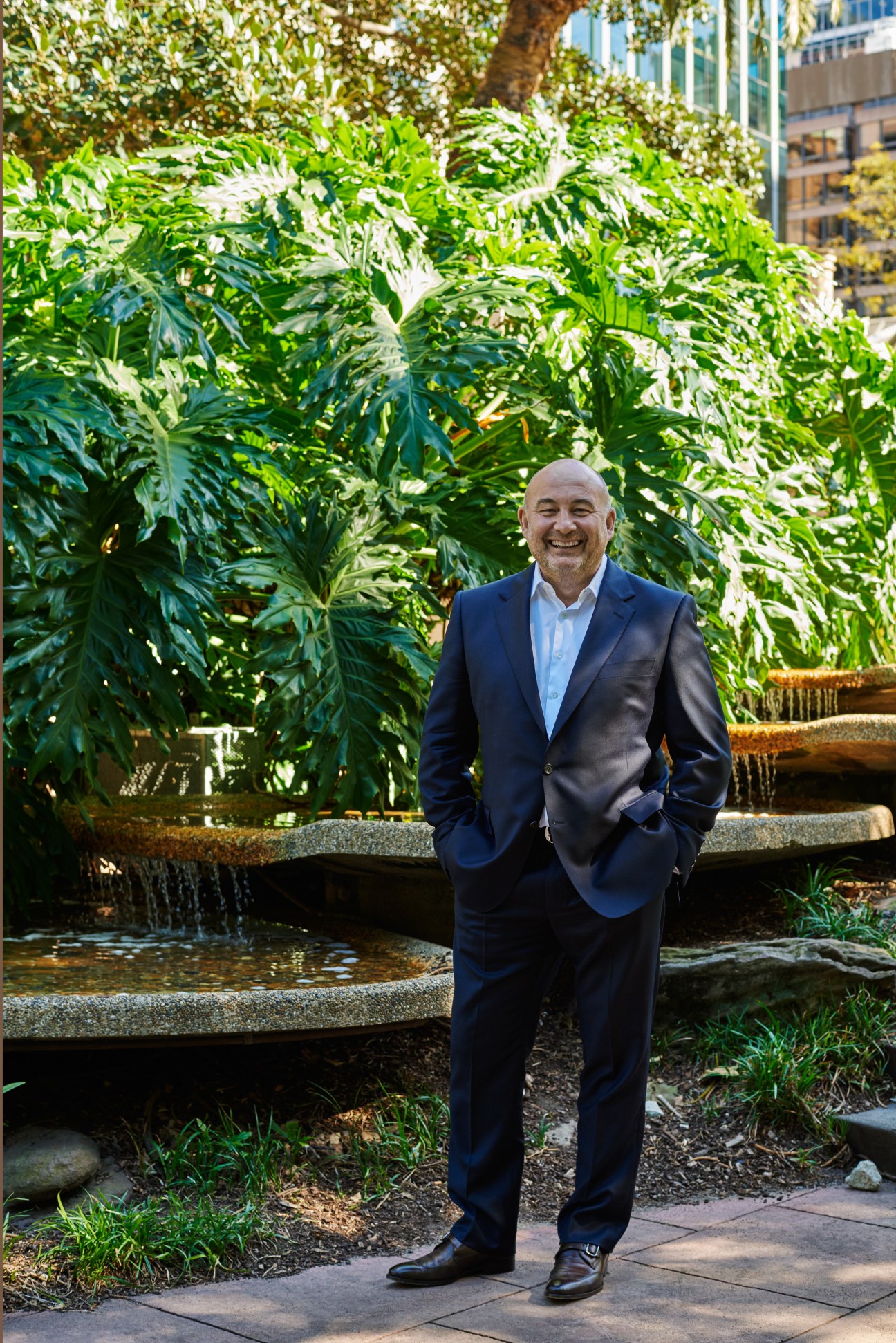 Man in navy blazer and light blue shirt standing in front of lush green foliage and water feature.