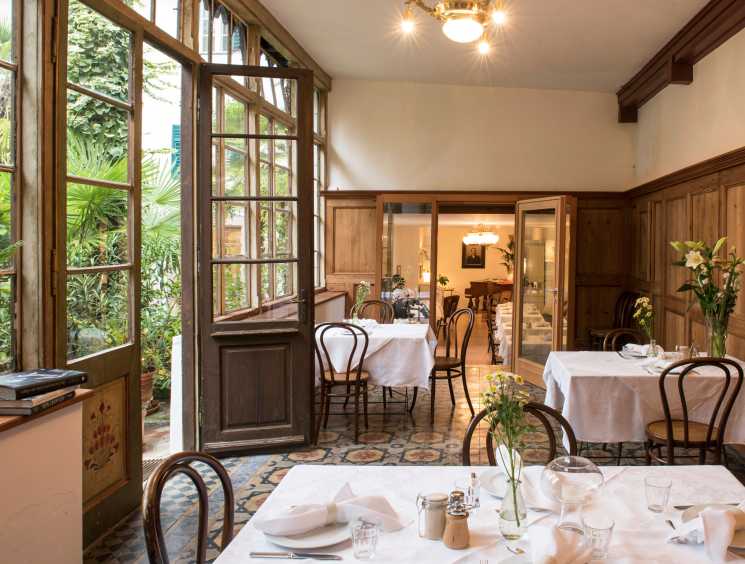 Fine dining restaurant interior with wooden paneling, white tablecloths, bentwood chairs, fresh flowers, and garden views through open doors.