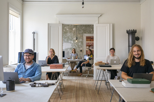 Open office workspace with wooden flooring, multiple staff members at desks, and wooden structural elements creating meeting pods.