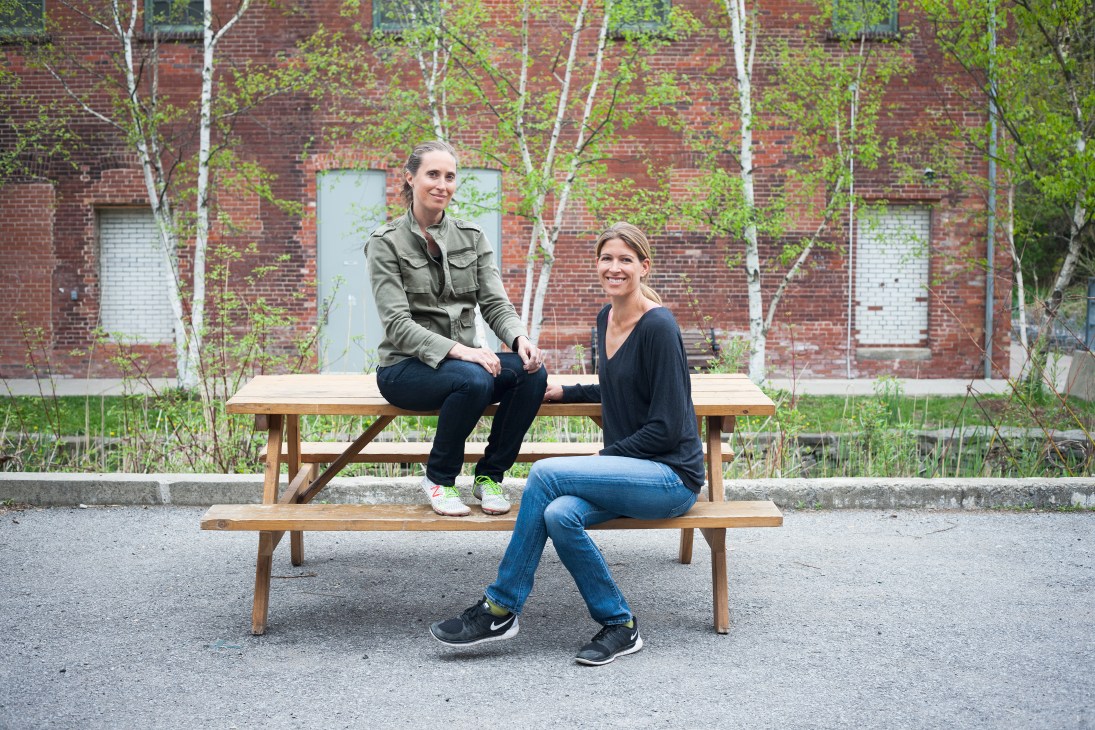 Two women sit on a wooden bench in front of a brick building with birch trees at Evergreen Brick Works in Toronto.