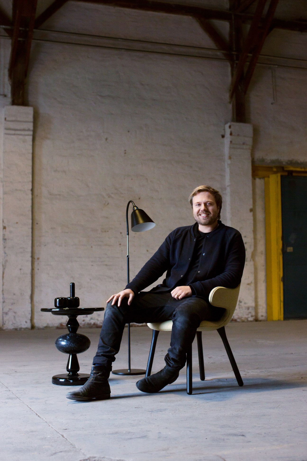 Klaus Kastbjerg sits in a modern chair next to a black pedestal table with an arc lamp in an industrial warehouse space.