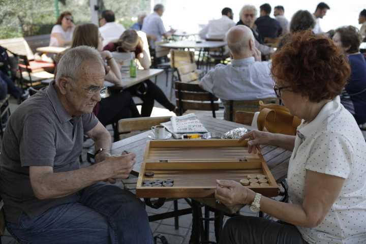 Two people playing backgammon at an outdoor café table with other patrons visible in the background.