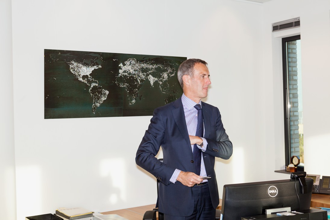 Man in dark suit standing in office with world map visualization on wall behind him.