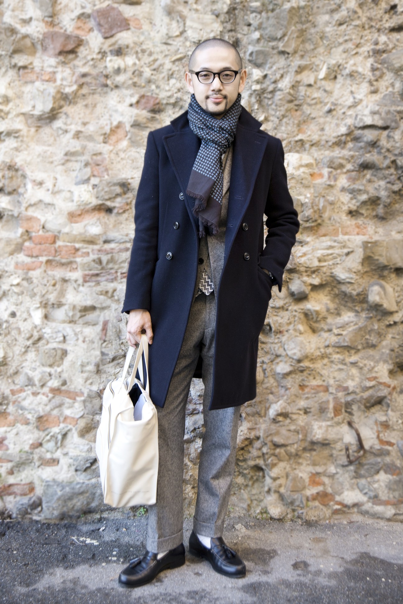 Man wearing navy coat, patterned scarf and glasses holding a white bag against a brick wall.