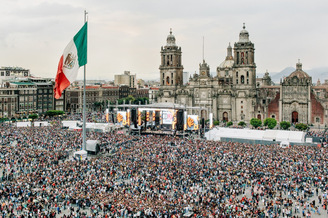 Large crowd gathered in Mexico City's Zócalo main square with concert stage and cathedral in background.