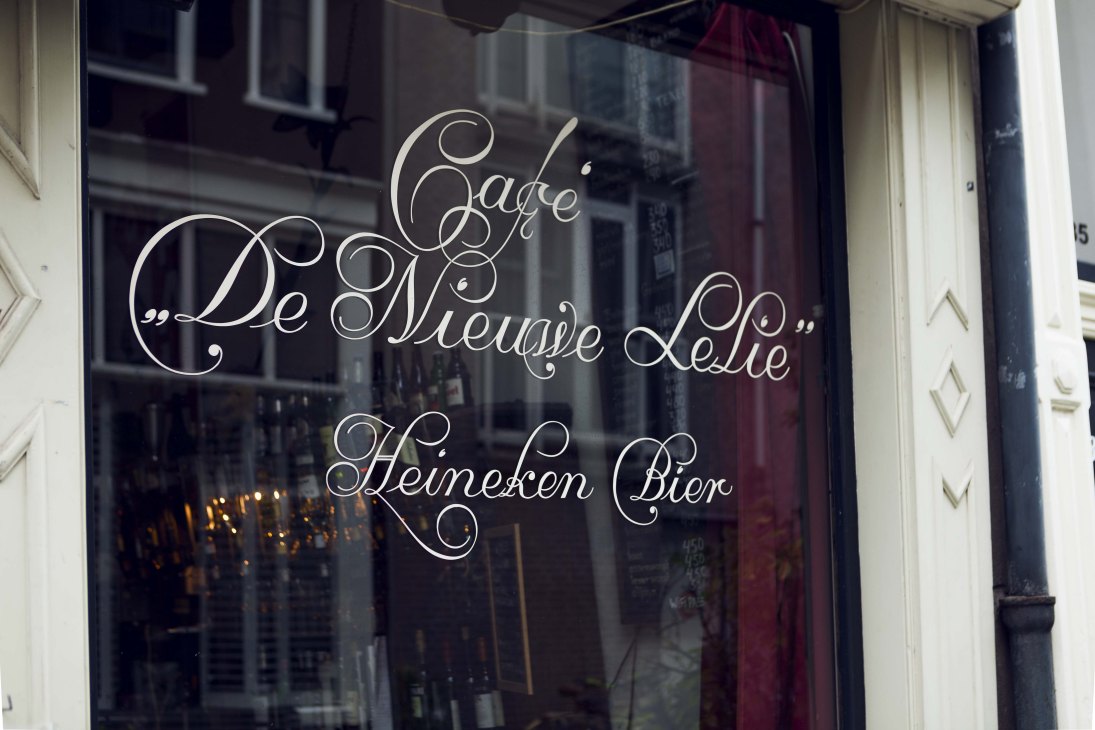 Hand-painted cursive lettering on a dark glass window reading "De Nieuwe Lelie" and "Heineken Bier"
