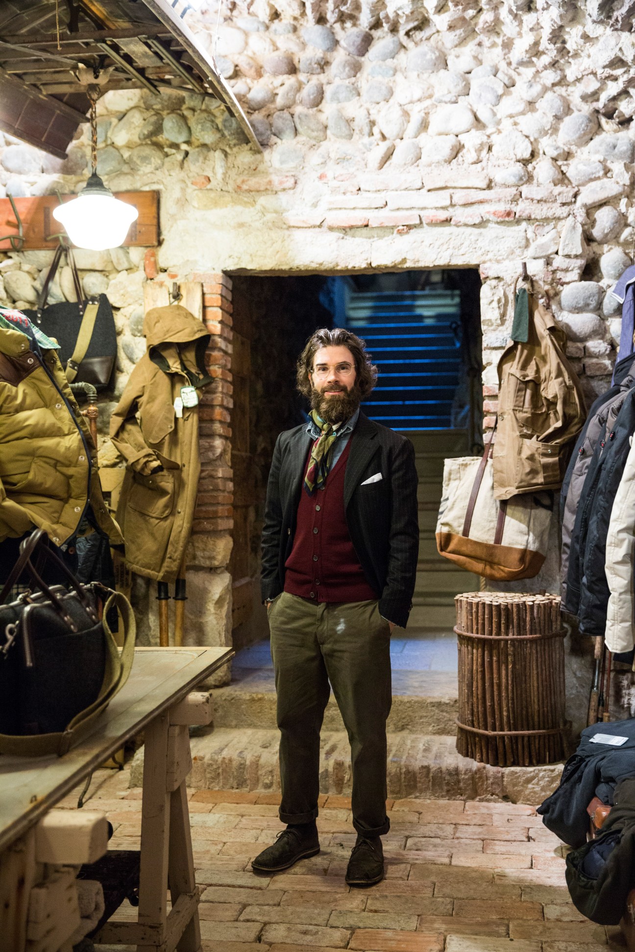 Man standing in menswear shop surrounded by hanging coats and outerwear against white brick walls.