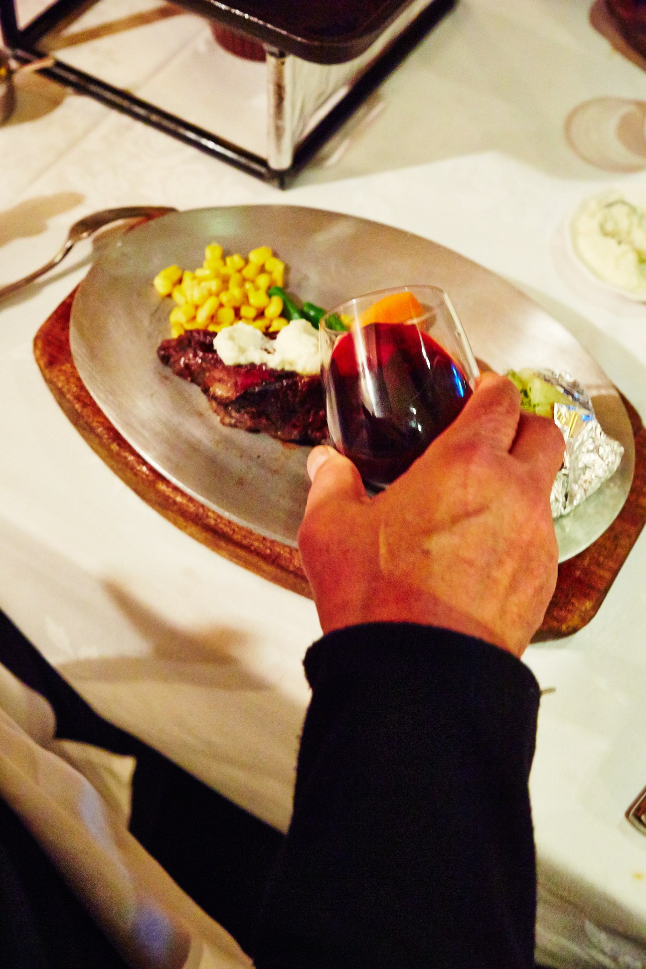 A hand holding a glass of red wine above a plate of rare steak with corn and white sauce.