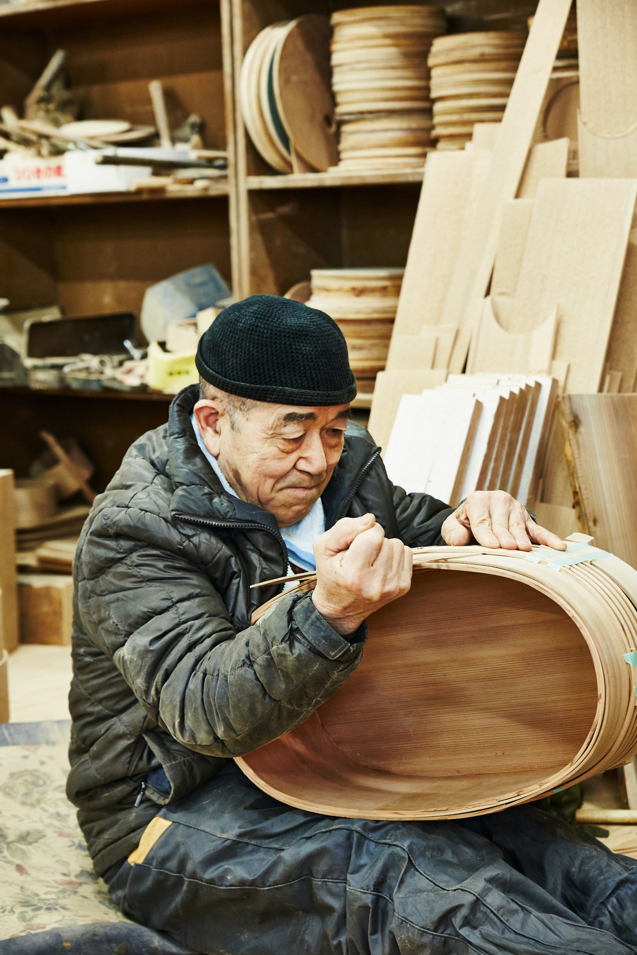 Elderly craftsman working on a bent cedar wood container in a workshop filled with wooden crafts and materials.