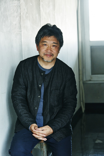 Japanese film director Hirokazu Koreeda sitting against a white wall wearing a dark jacket.
