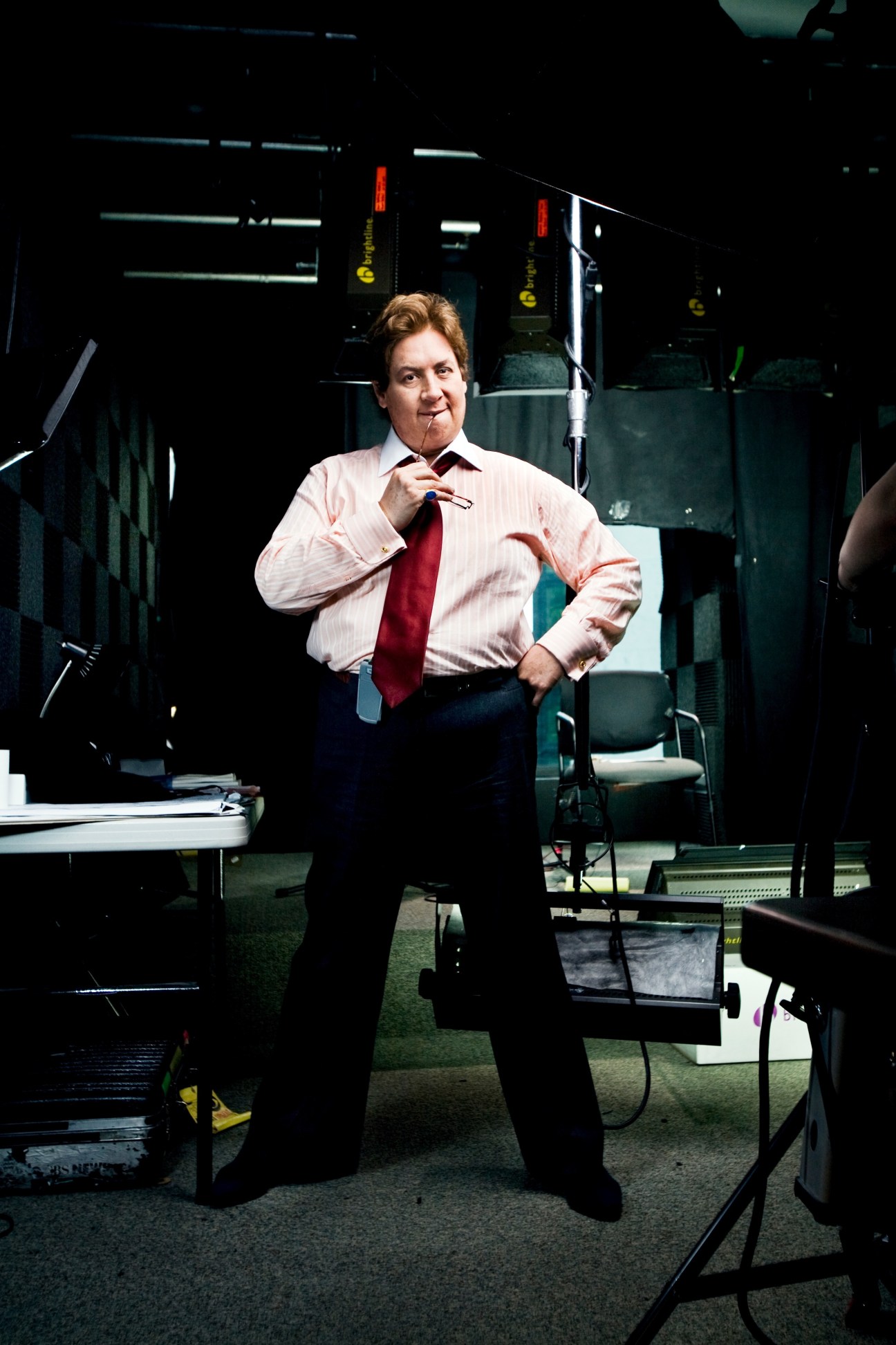 Man in pink shirt and red tie standing in a television studio holding a microphone.