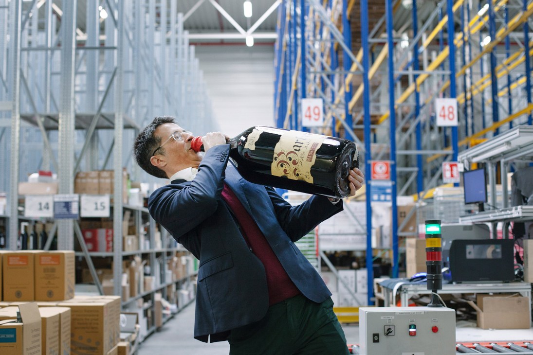 Man in business attire drinks from a wine bottle in a warehouse with shelving and numbered racks.