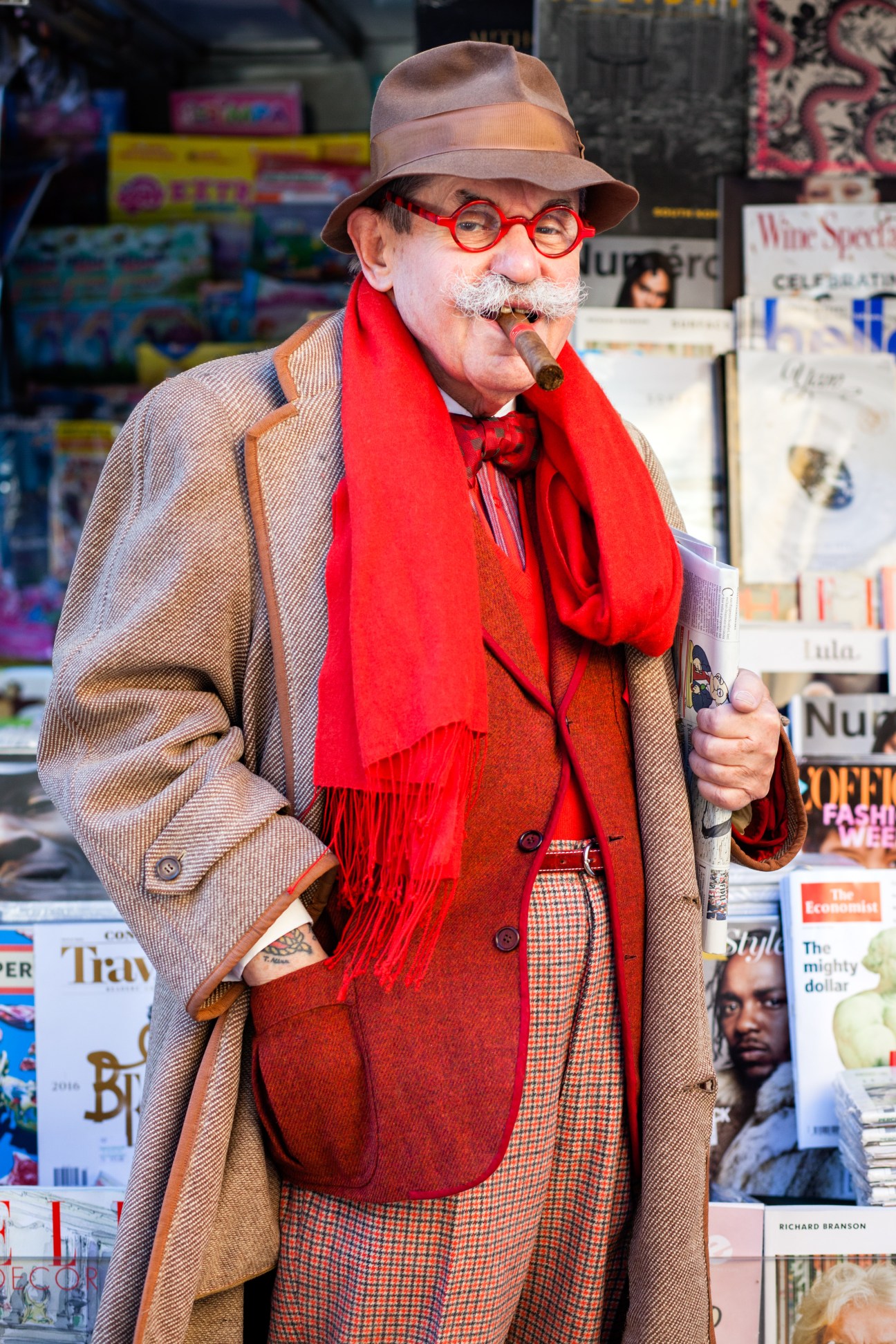 Man in beige blazer and red scarf with red glasses smoking cigar near newsstand display