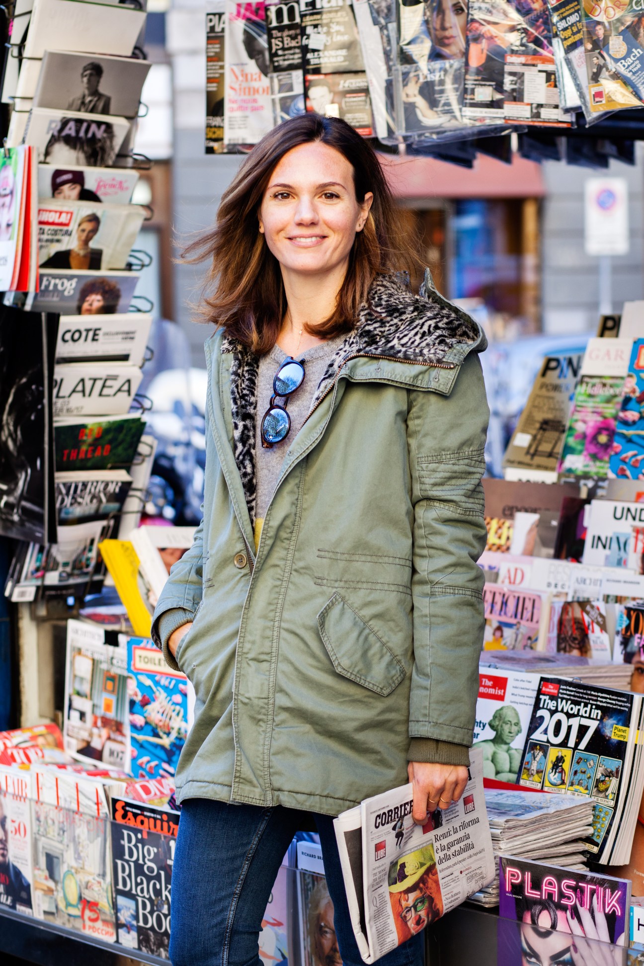 Woman standing at a newsstand holding a newspaper surrounded by magazines and publications.