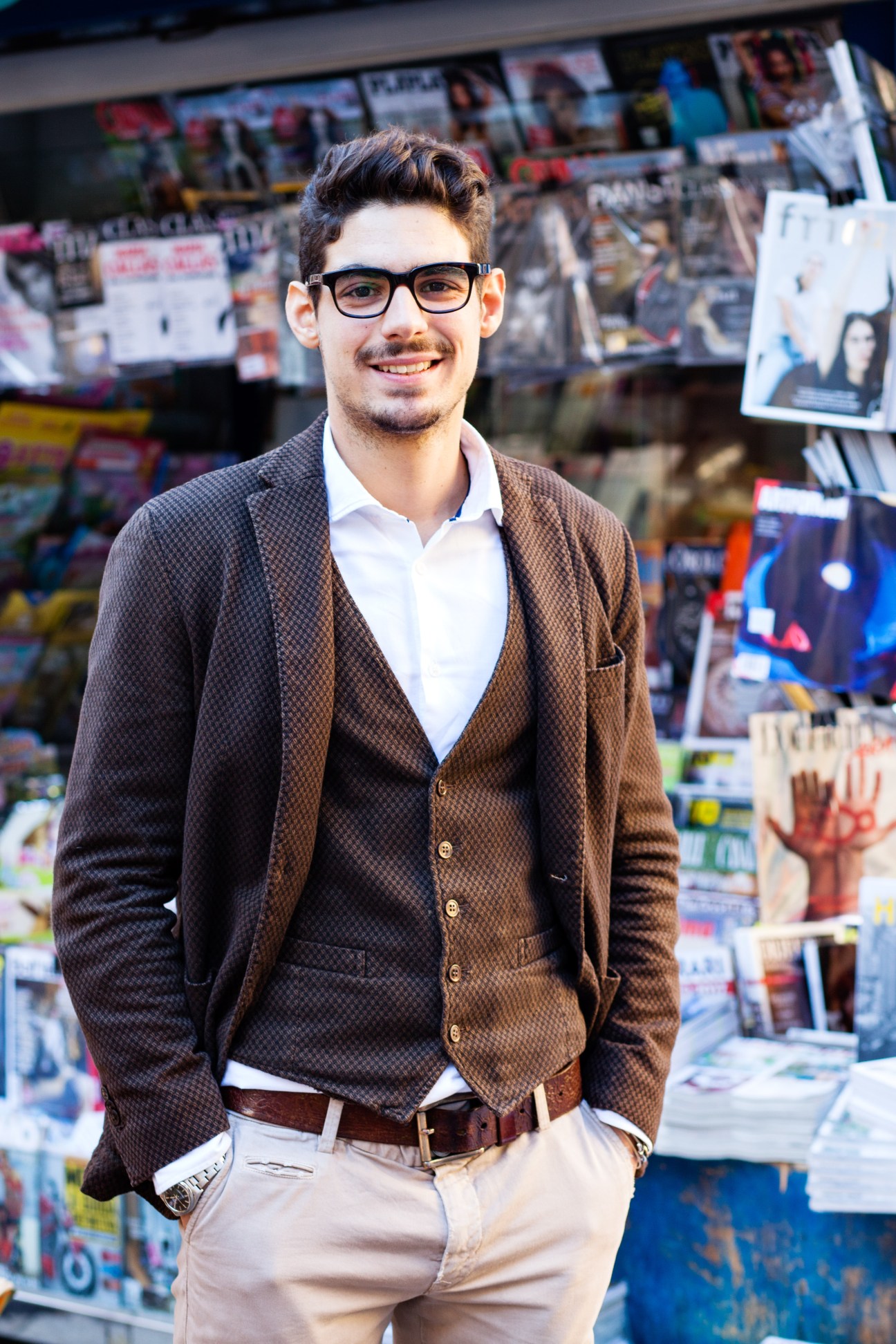 Young man with glasses smiling in front of a news kiosk displaying various newspapers and magazines.