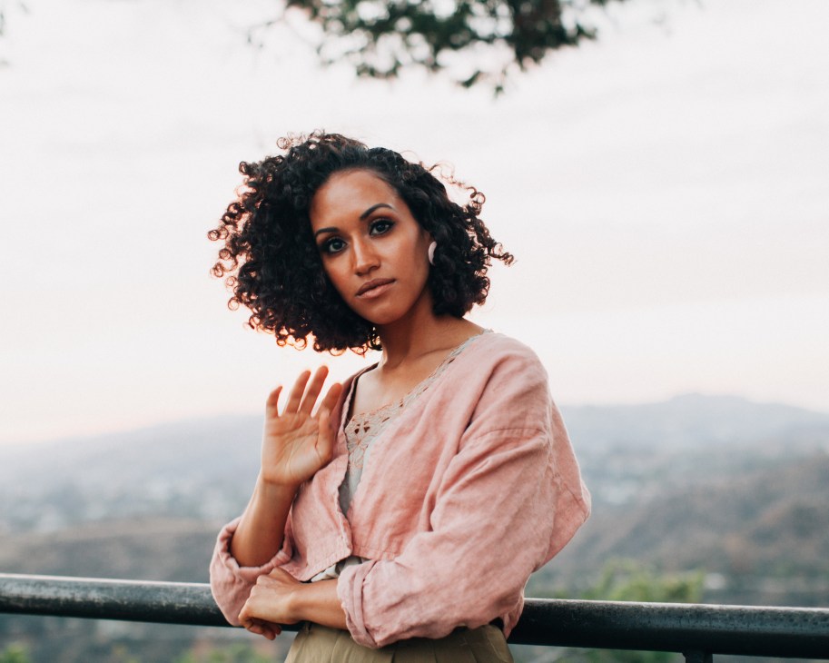 Musician Kadhja Bonet poses outdoors with curled hair, wearing a pink linen shirt against a blurred hillside background.