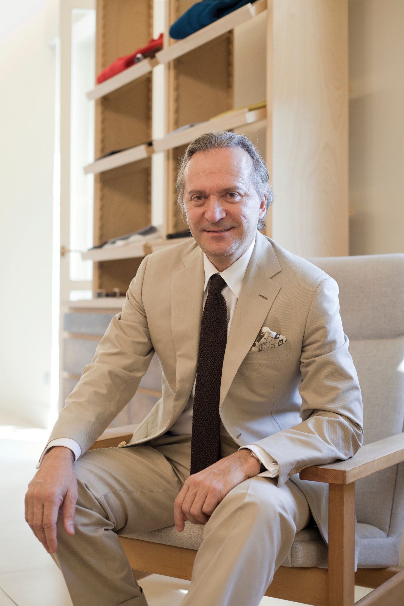 Man in beige suit and dark tie sitting on wooden chair with leather goods displayed on shelves behind him.