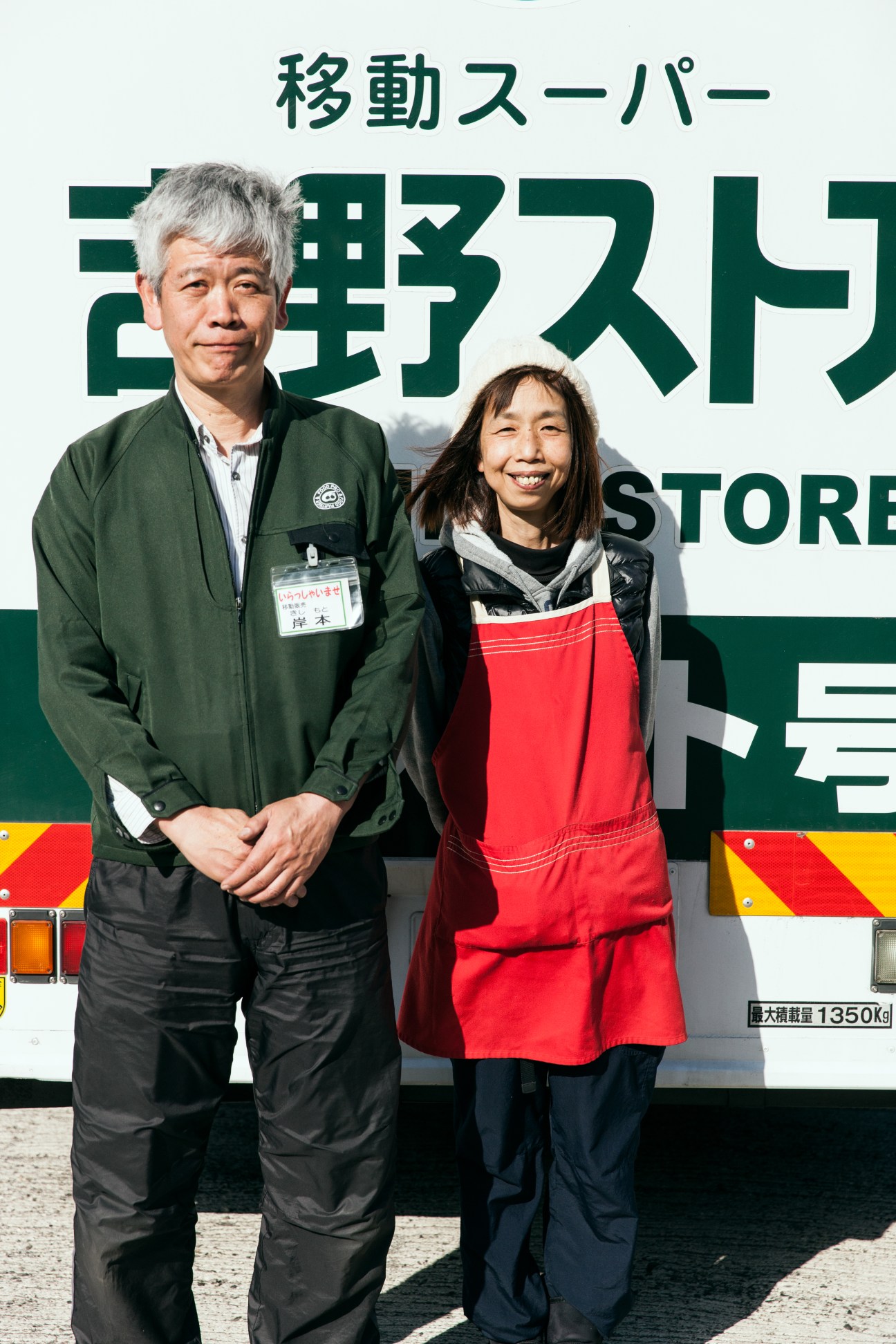 Two Yoshino Store employees stand in front of their mobile grocery truck with Japanese signage.