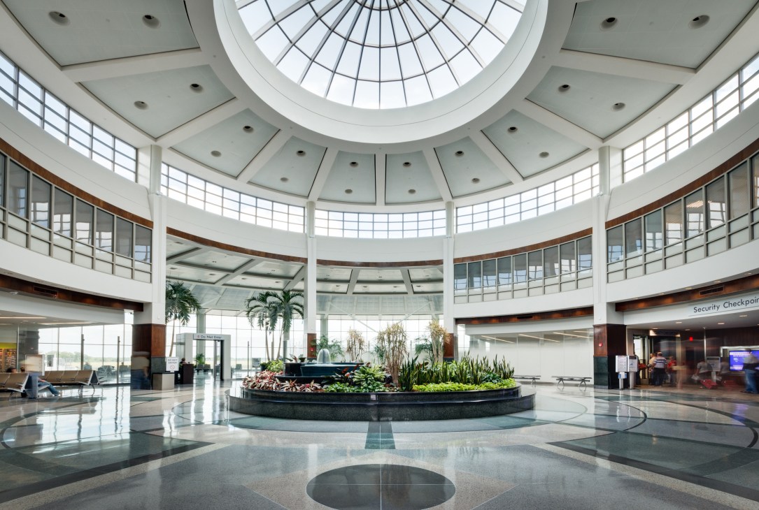 Modern airport terminal interior with circular skylight, plants, and curved railings.