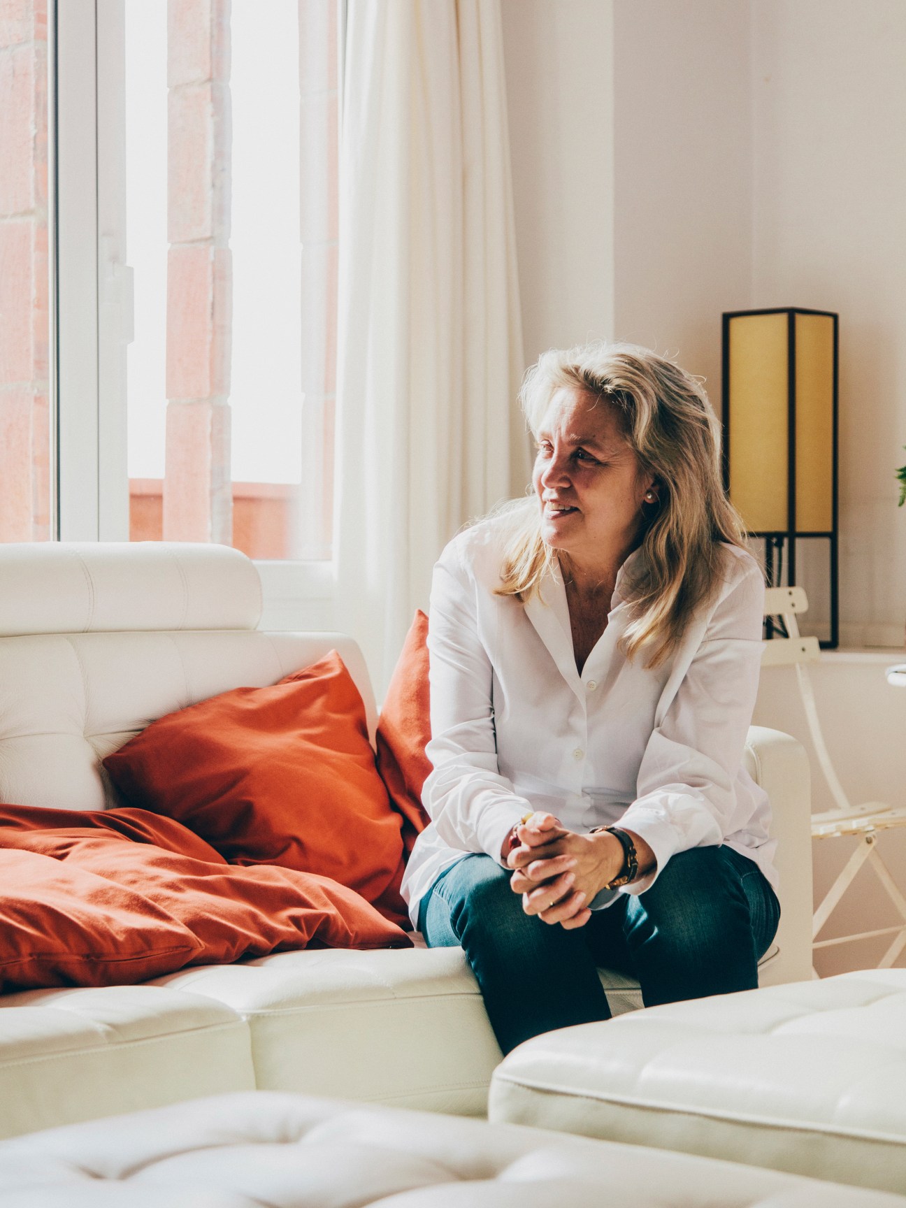Woman sitting on white sofa with orange cushions in a bright apartment with terracotta-colored building visible through window.