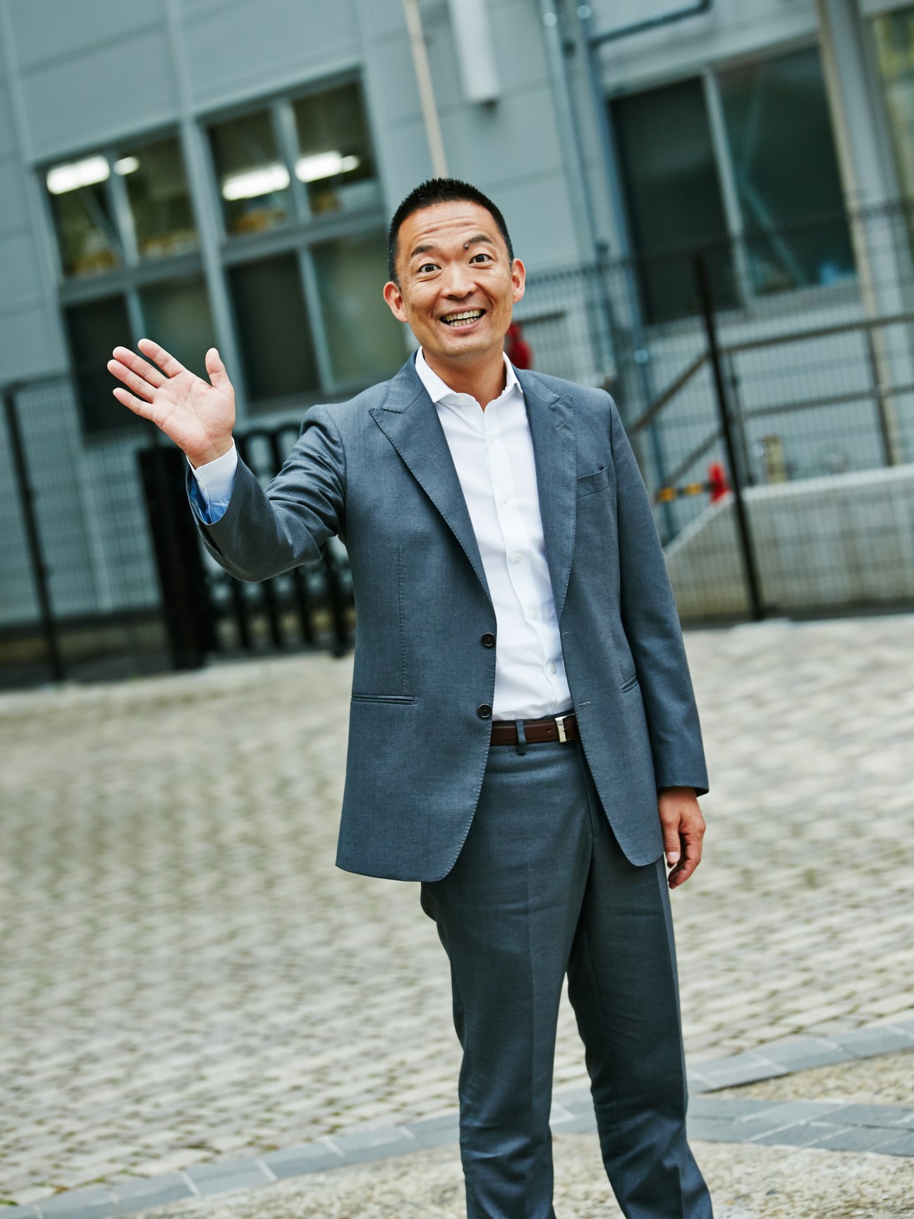 Ken Hasebe, Mayor of Shibuya, wearing a grey blazer and white shirt, waving and smiling in front of a modern glass building.