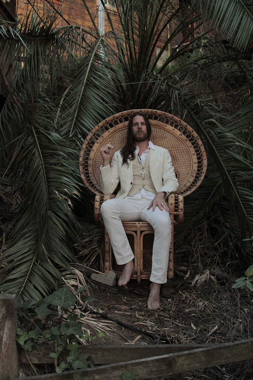 Man with long dark hair in cream suit seated on wicker peacock chair surrounded by tropical plants.