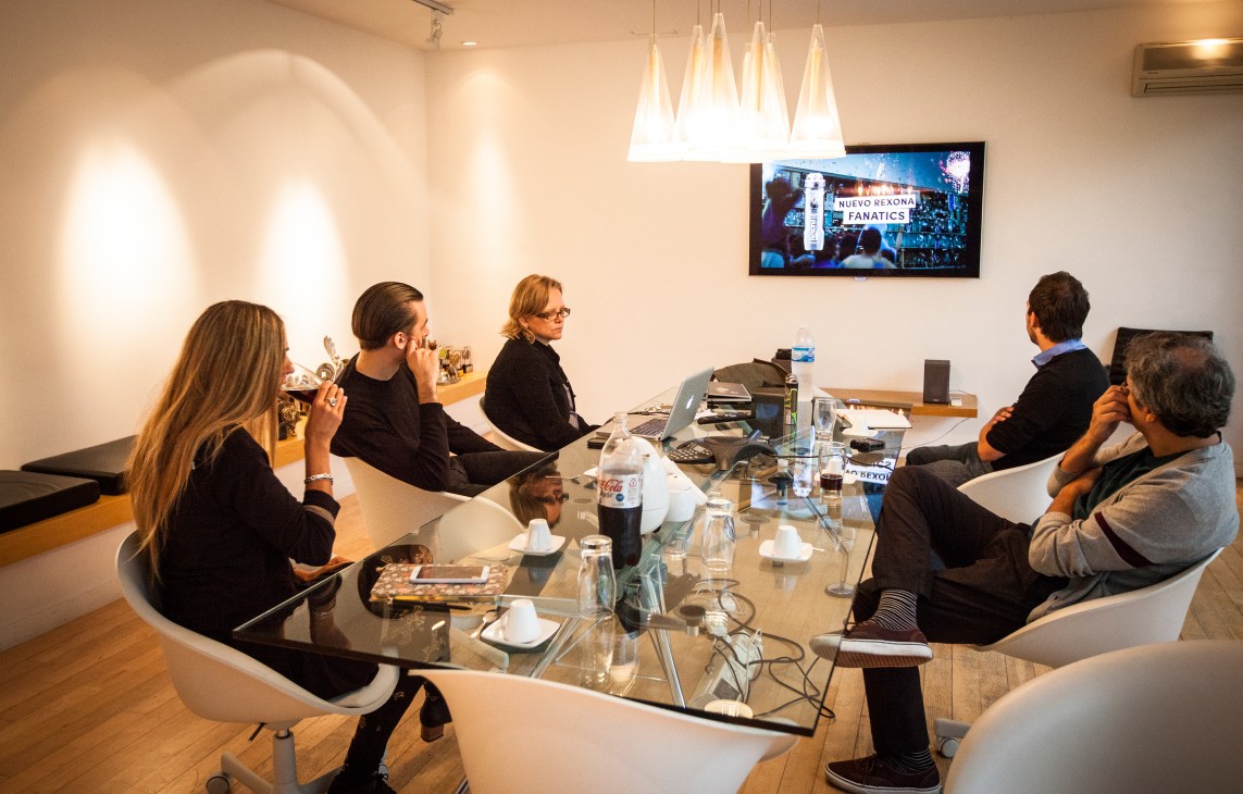 People seated around a glass conference table in a modern office meeting room with a TV screen displaying a Rexona Fanatics advertisement.