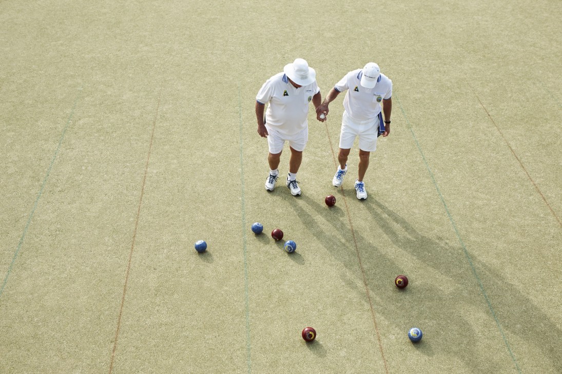 Two lawn bowls players in white uniforms on a manicured bowling green with bowls scattered across the lawn.