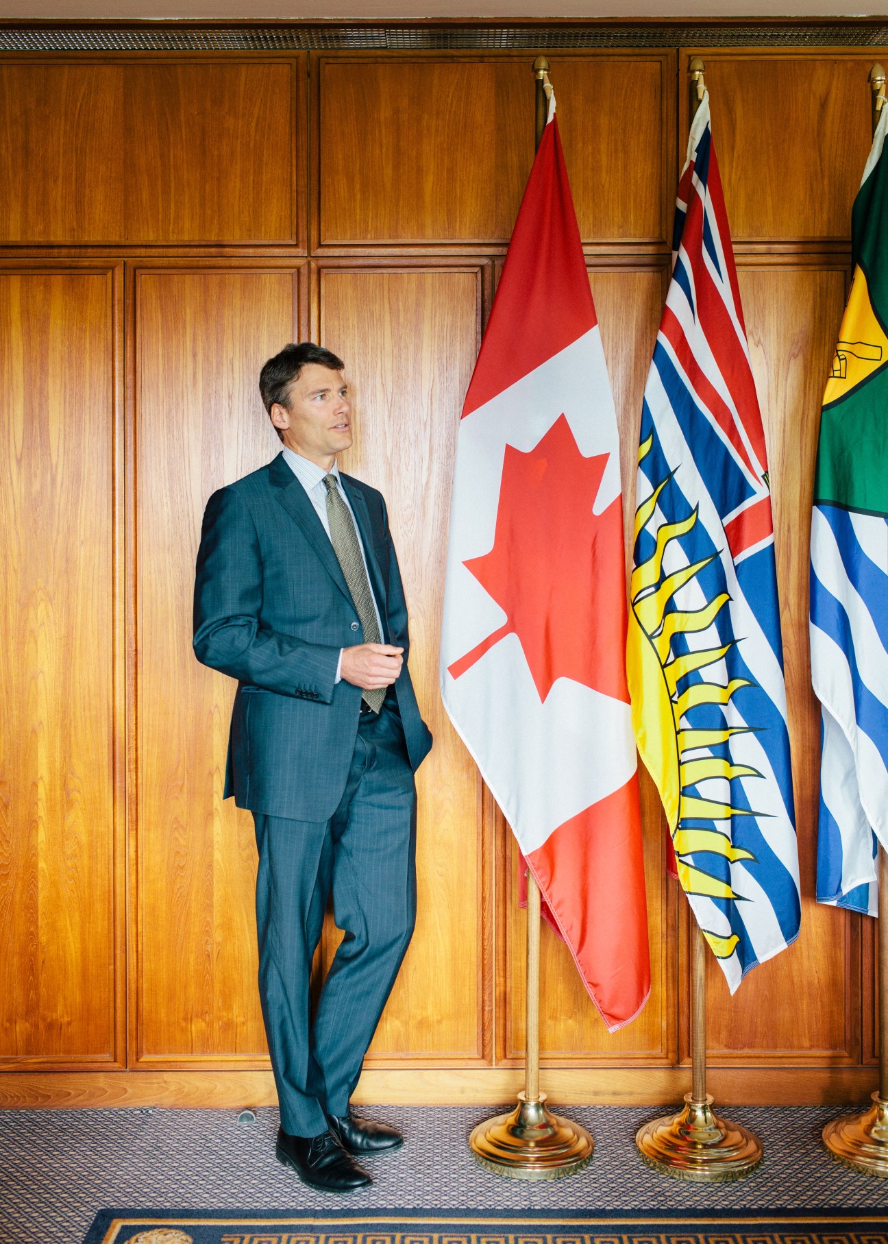 Man in dark suit standing against wooden paneling with Canadian and British Columbia flags.