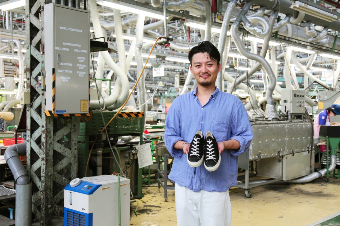 Kenta Matsunaga holds canvas-and-rubber Moonstar trainers in a Japanese shoe factory with industrial equipment in the background.