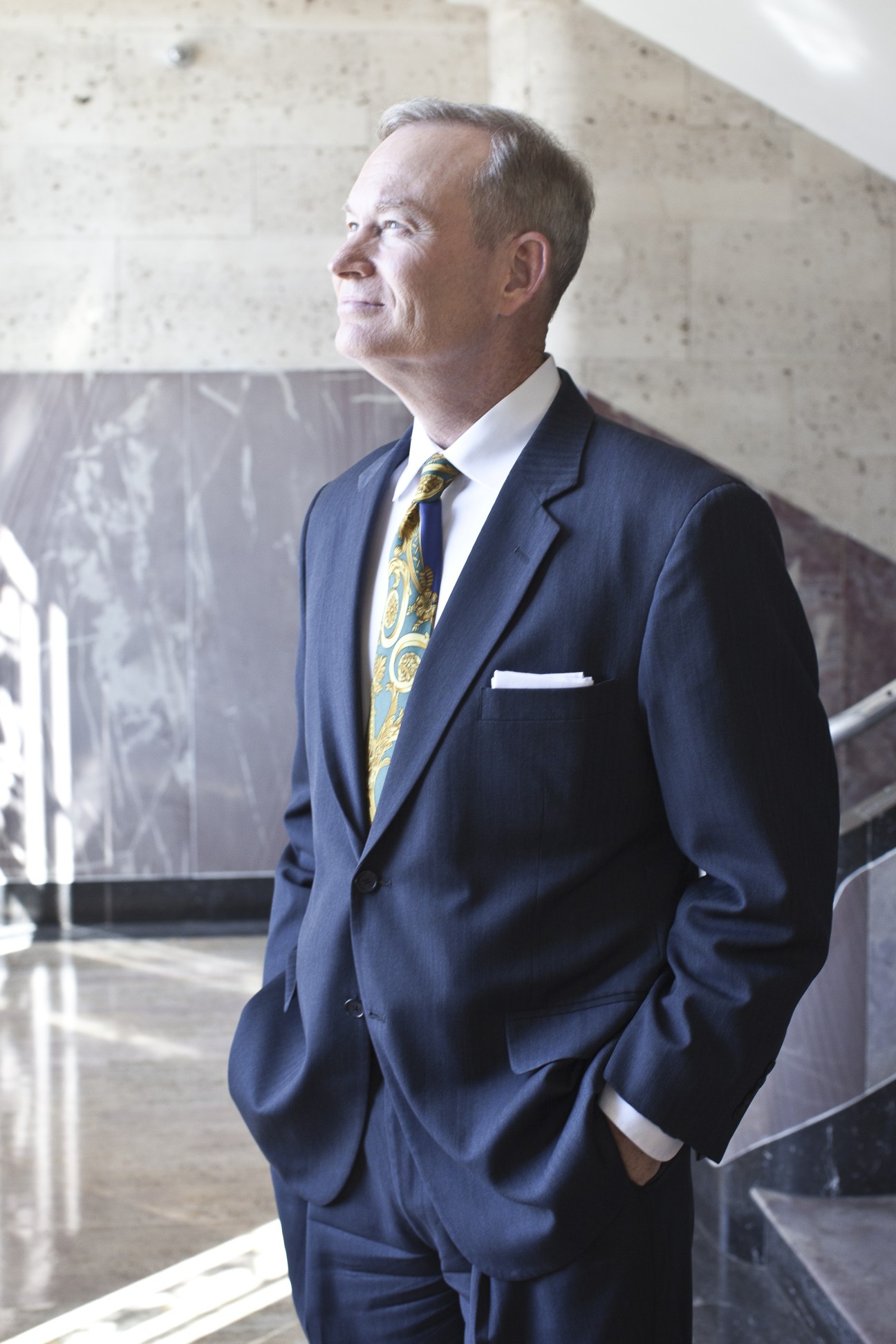 Man in navy suit and patterned tie standing in modern building corridor.