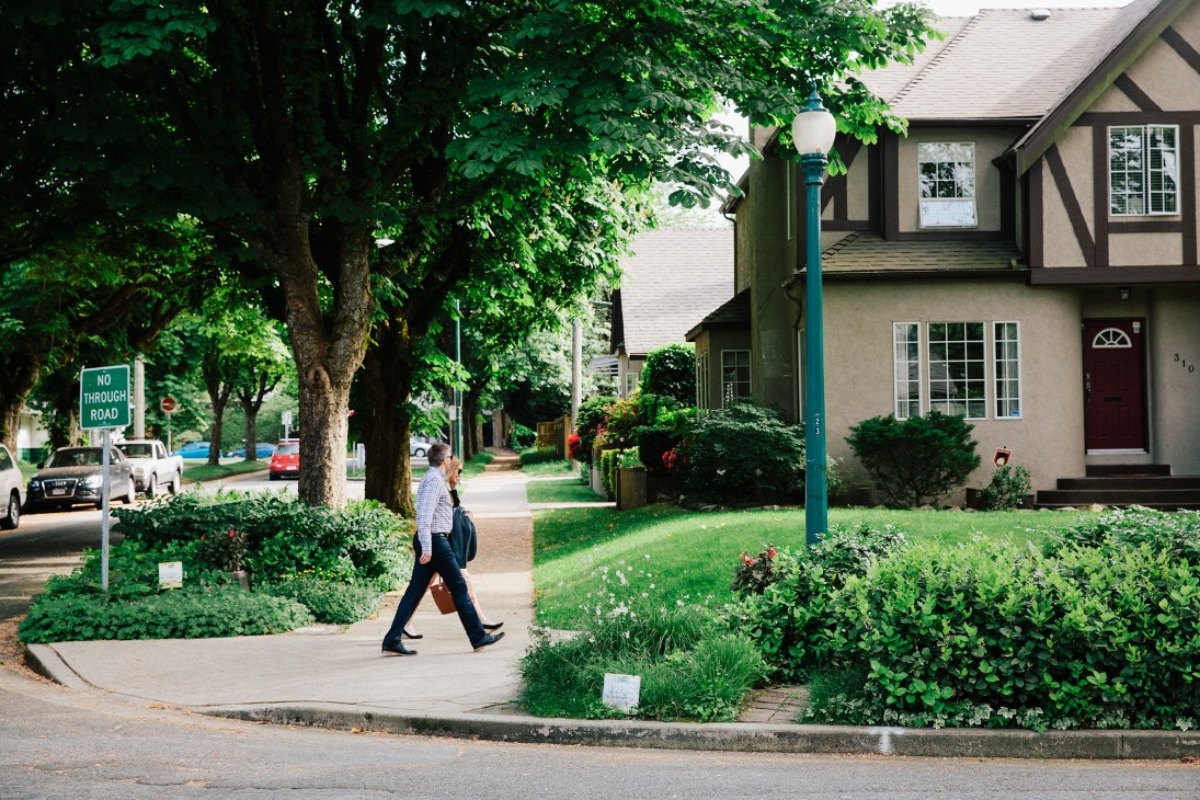 Person walking past mature trees and landscaped gardens on a residential street in Vancouver.