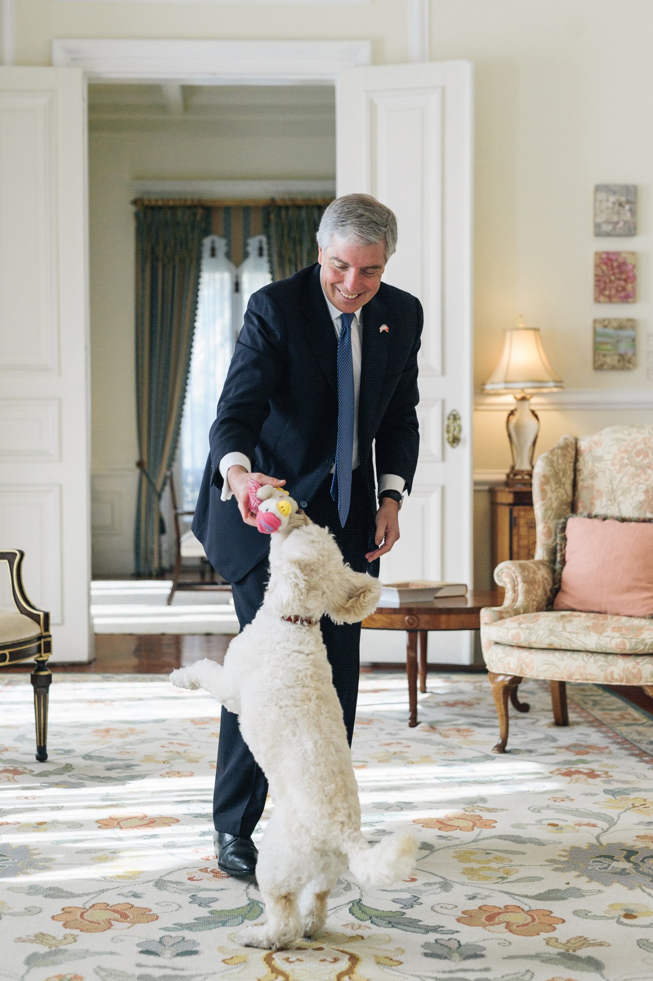 Man in dark suit plays with a white dog standing on hind legs indoors.