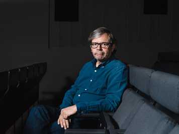 Pedro Borges, a man wearing glasses and a blue shirt, sits on a grey couch in a dark room.
