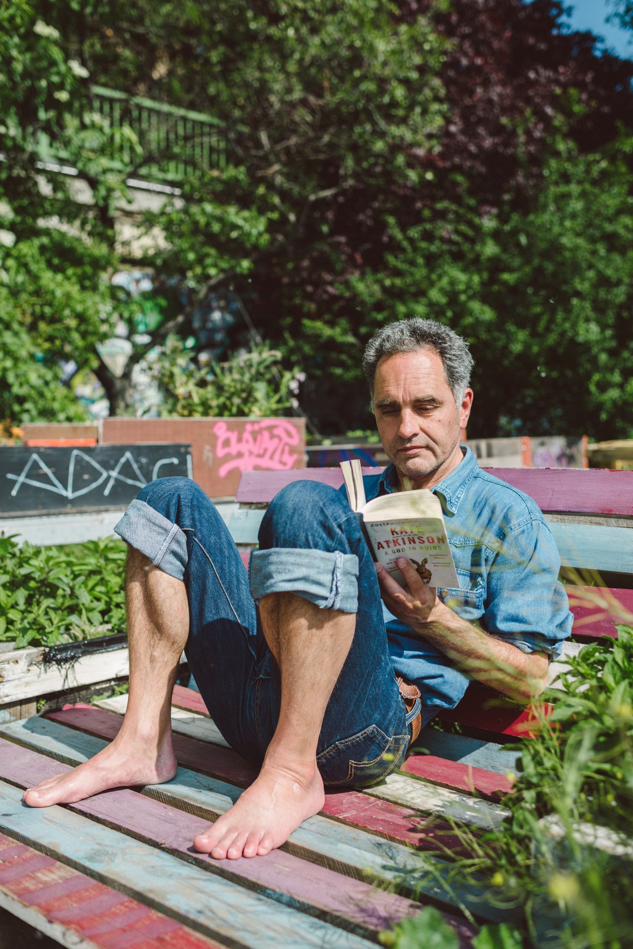 Man reading a book barefoot on a colorful wooden deck surrounded by green foliage and ivy-covered walls.