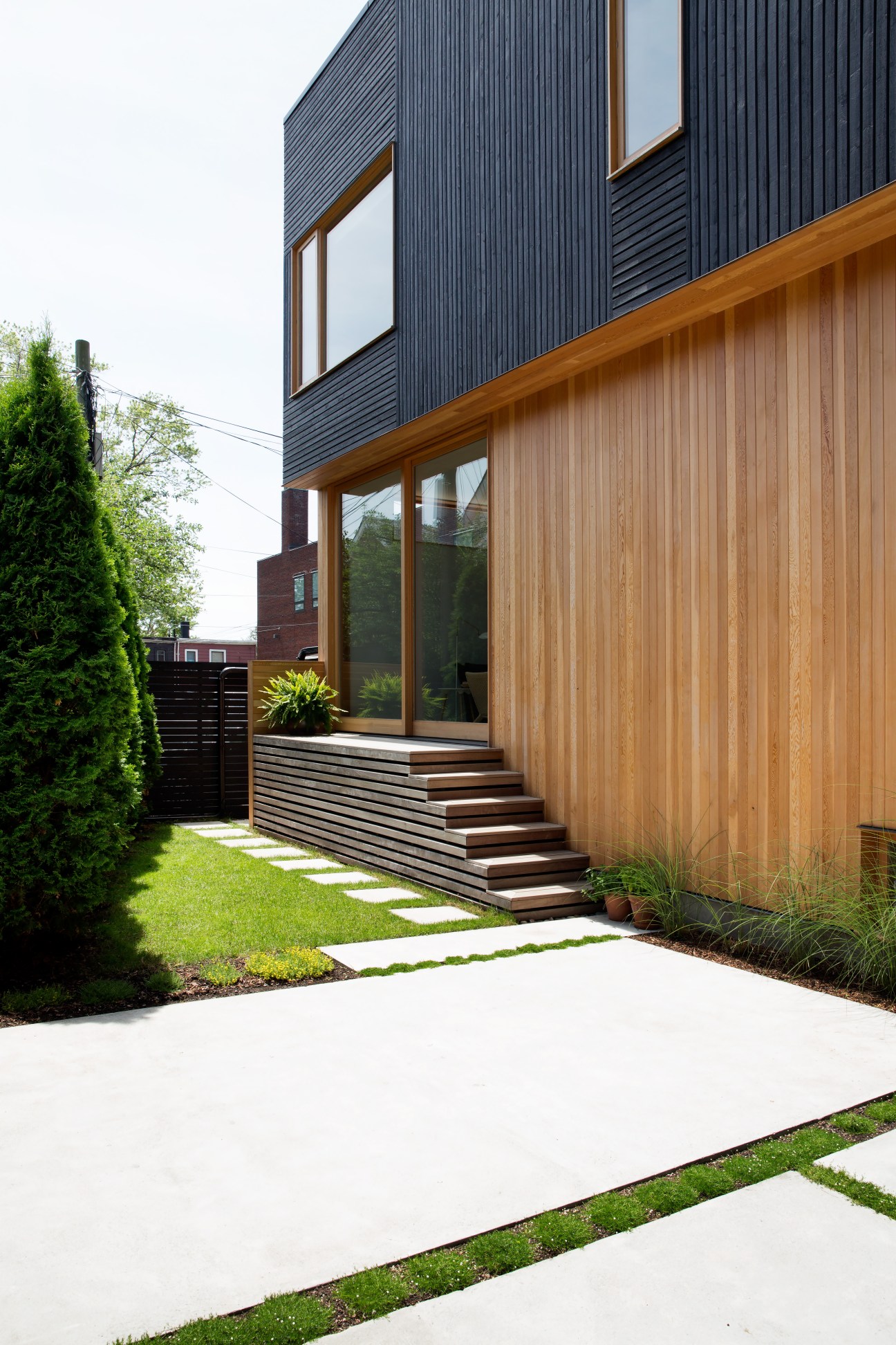 Modern wooden house with black upper cladding and natural wood lower facade, featuring large windows and wooden steps leading to entrance.
