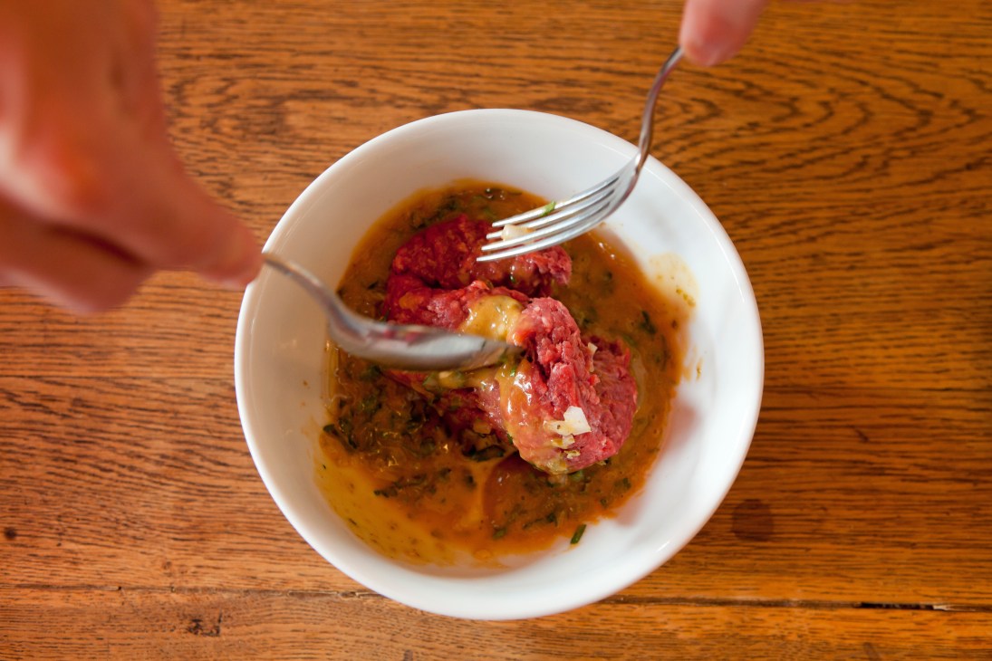 A fork mixes raw steak tartare with sauce in a white bowl on a wooden table.