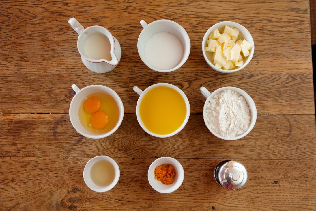 Crêpes Suzette ingredients arranged in white bowls on a wooden table, including milk, sugar, butter, eggs, orange juice, flour, and Grand Marnier.