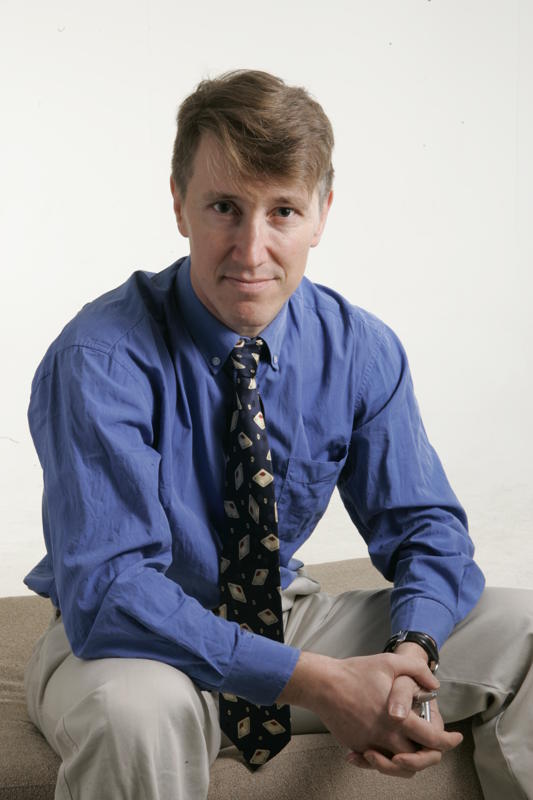 Portrait of Andrew Salmon, a writer in blue shirt and patterned tie, seated against white background.