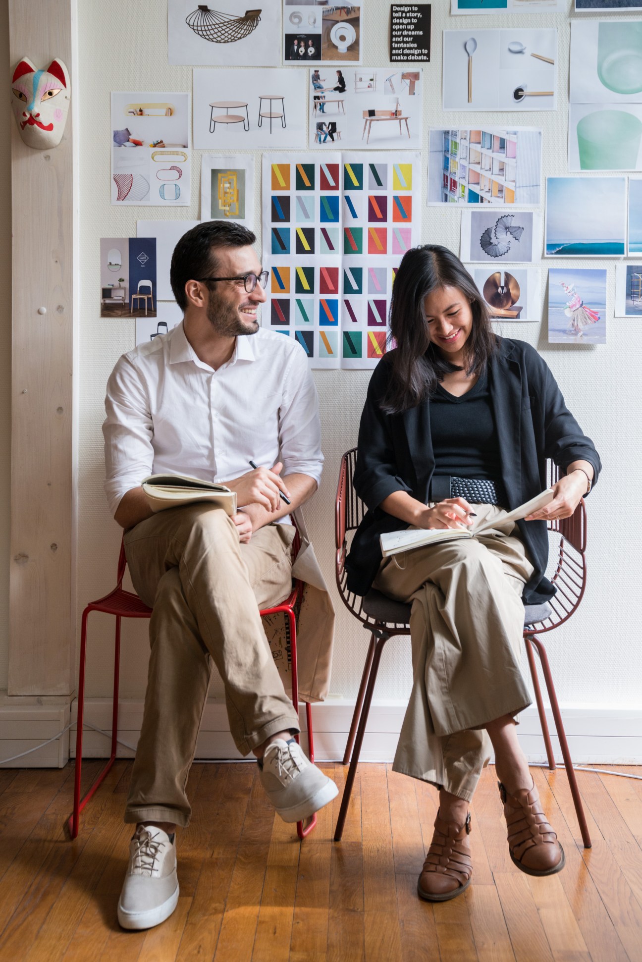 Two designers sitting on red wire chairs reviewing portfolios in a studio with design mood boards on the wall