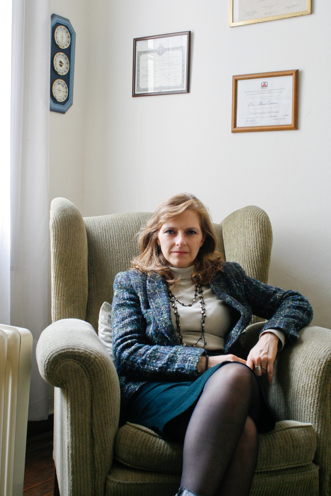 Psychoanalyst Alicia Costanzo Aldecosea seated in a beige armchair in her office with framed certificates on the wall behind her.