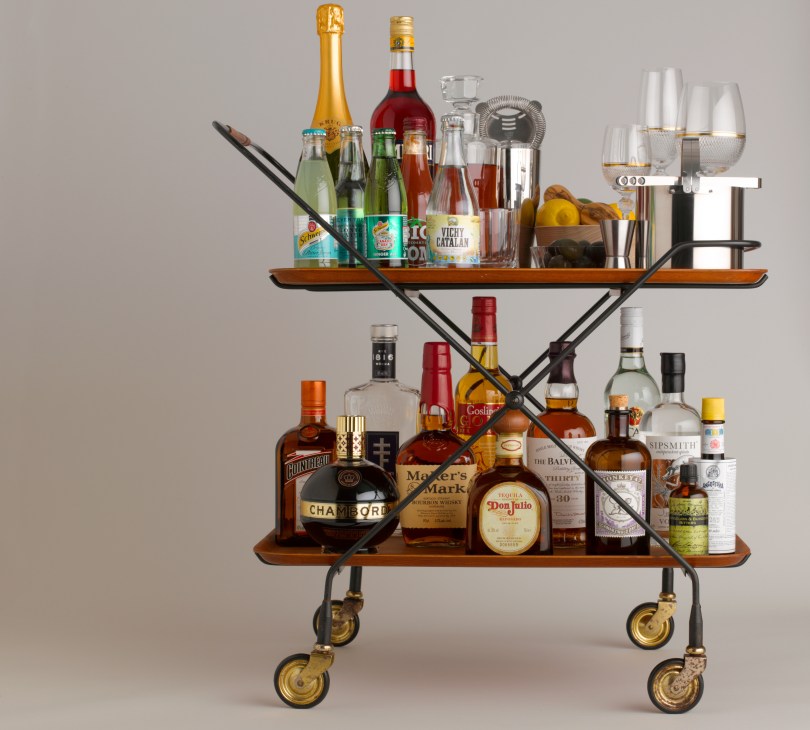 A well-stocked drinks trolley with two wooden shelves displaying spirits, mixers, glassware, and cocktail tools.